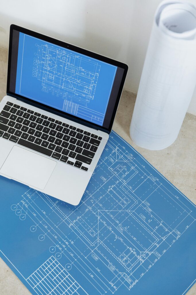 Laptop showing engineering blueprint with rolled paper and drawings on table.