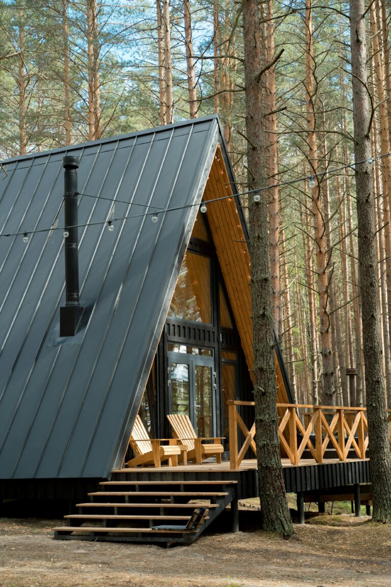A modern A-frame cabin nestled among tall trees in a peaceful woodland, featuring a wooden porch and black metal roof.